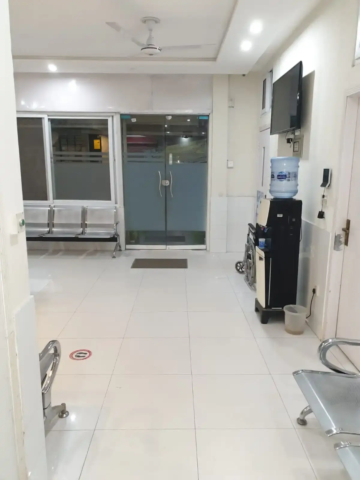 Glass-door entrance and lobby of Al Shifa Maternity Home with metal seating, a water cooler and a clean tiled floor.