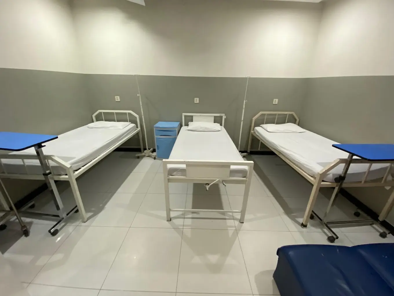 Patient care area with three single beds made with clean white linen, a bedside cabinet and a tiled floor.