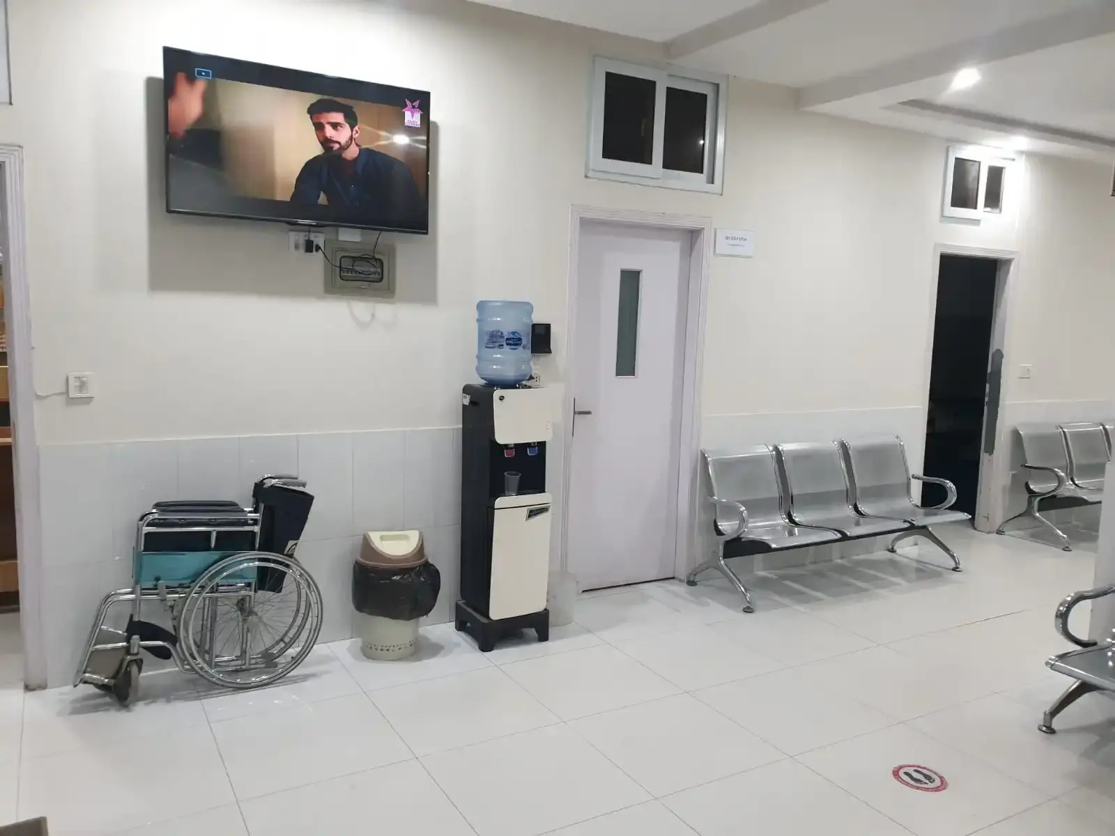 Waiting lounge with rows of seating, a wall-mounted television, a water cooler and a wheelchair.